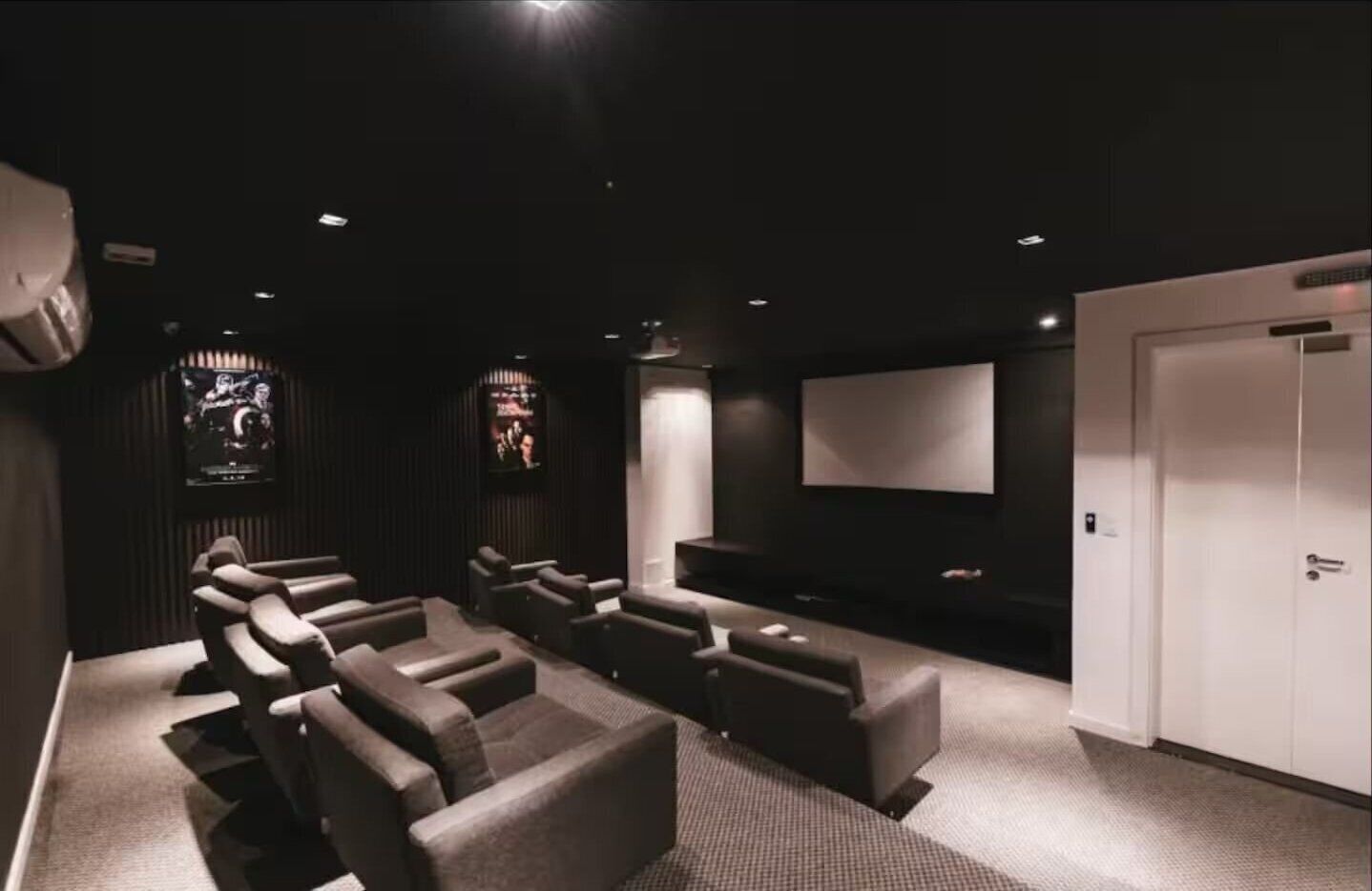 Een filmzaal in donkere kleuren met meerdere rijen stoelen, een groot scherm en een deur.