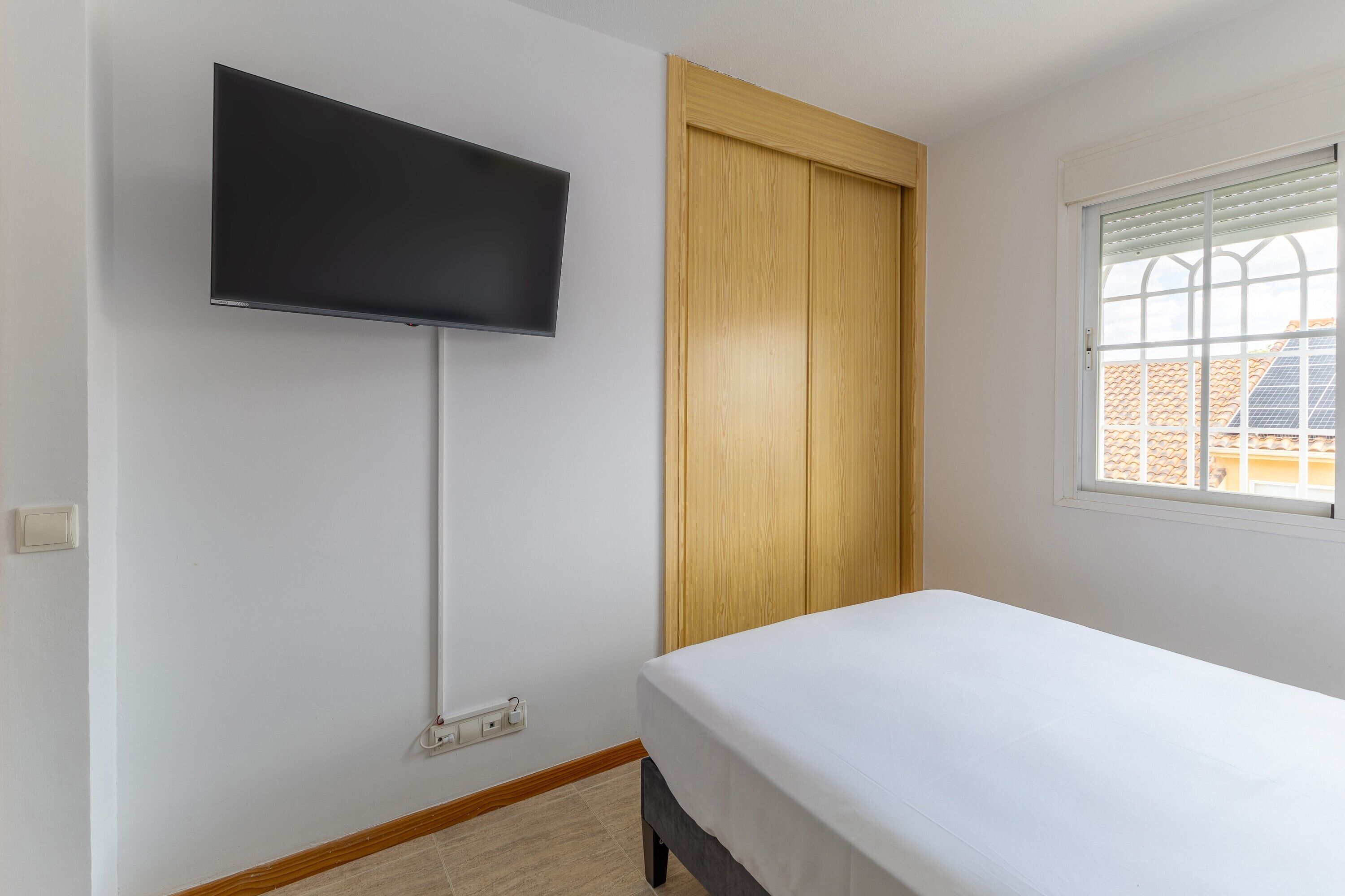 Una habitación con cama, televisor montado en la pared, armario de madera y ventana con persianas.