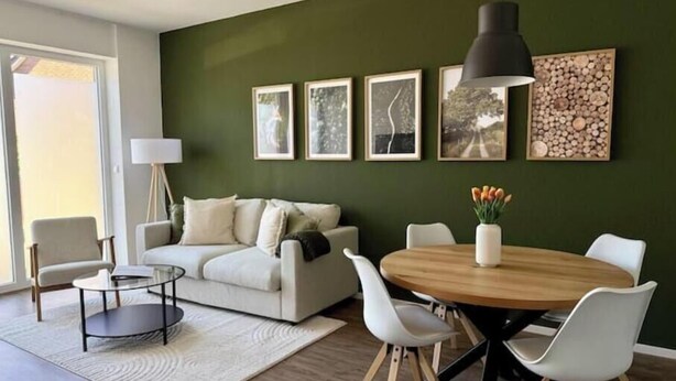 A modern living room with a green accent wall, a round wooden dining table, white chairs, a white sofa, and a floor lamp. - Modern comfort in the heart of Bocholt - Stylish apartment for up to 4 guests (Bocholt)