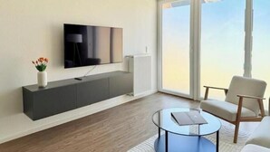 A modern living room with a flat-screen TV, a white radiator, a glass coffee table, and a large window with a view. - Modern comfort in the heart of Bocholt - Stylish apartment for up to 4 guests (Bocholt)