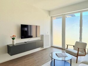 Ein modernes Wohnzimmer mit Flachbildfernseher, weißem Heizkörper, Glastisch und großem Fenster mit Aussicht.