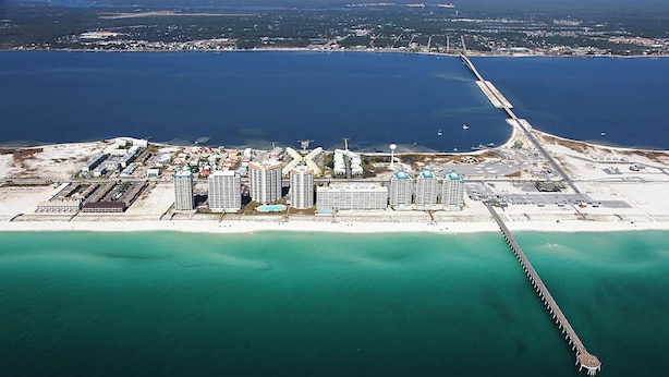 Vue aérienne d’une zone côtière comprenant un port de plaisance, plusieurs immeubles de grande hauteur et une longue jetée s’avançant dans l’eau.