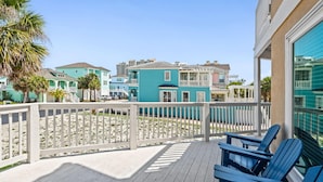 Une terrasse en bois avec des chaises bleues, une clôture blanche et, au fond, des bâtiments turquoise.