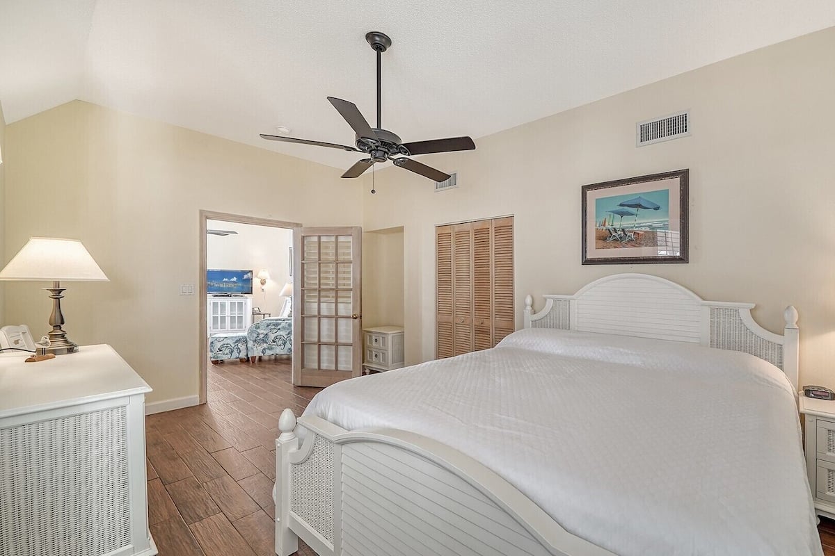 A bedroom with a bed, bedside lamps, a ceiling fan, a wardrobe, and a painting on the wall.