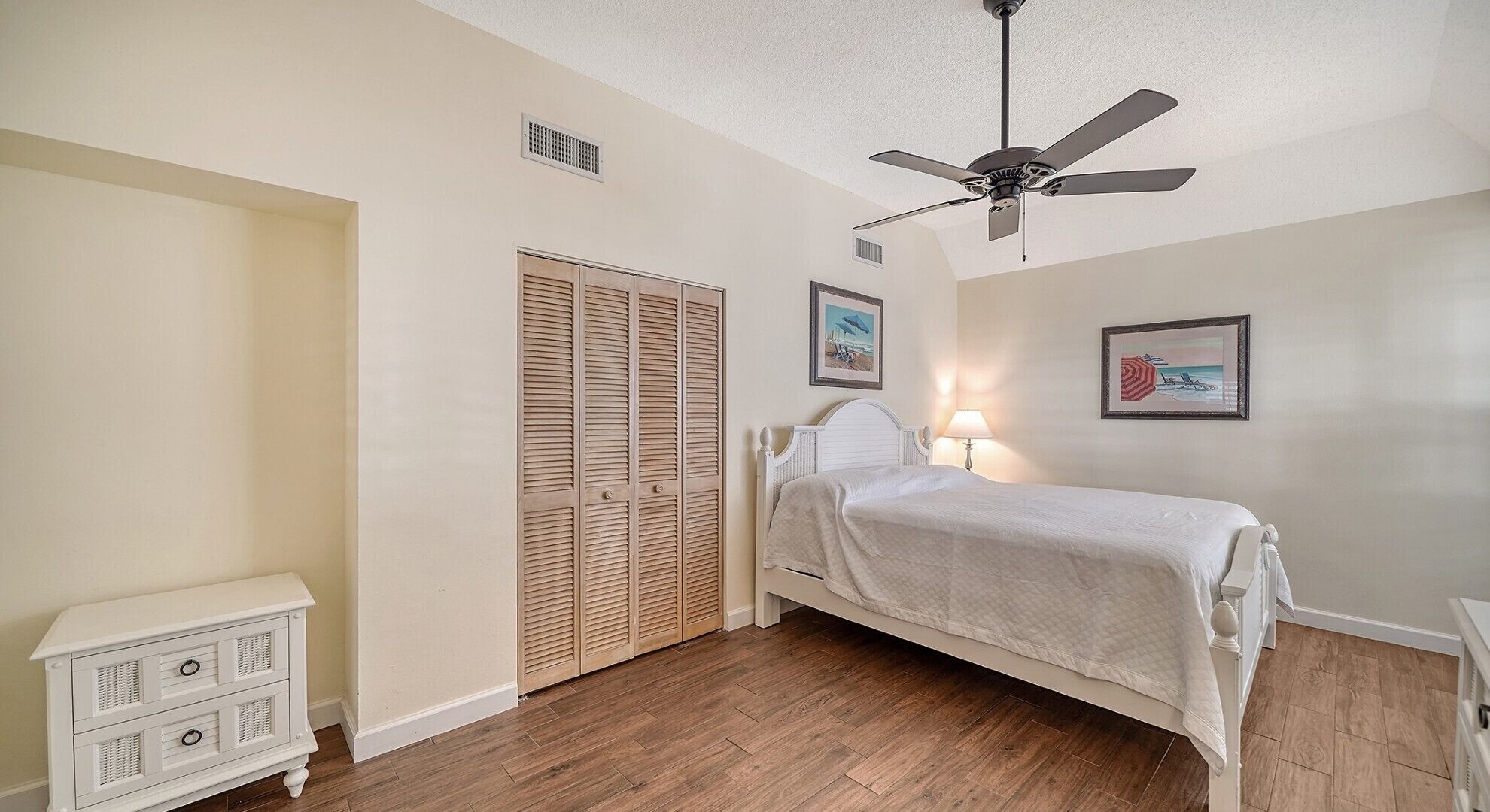 A bedroom with a bed, a nightstand, a ceiling fan, a wardrobe, and two framed pictures on the wall.