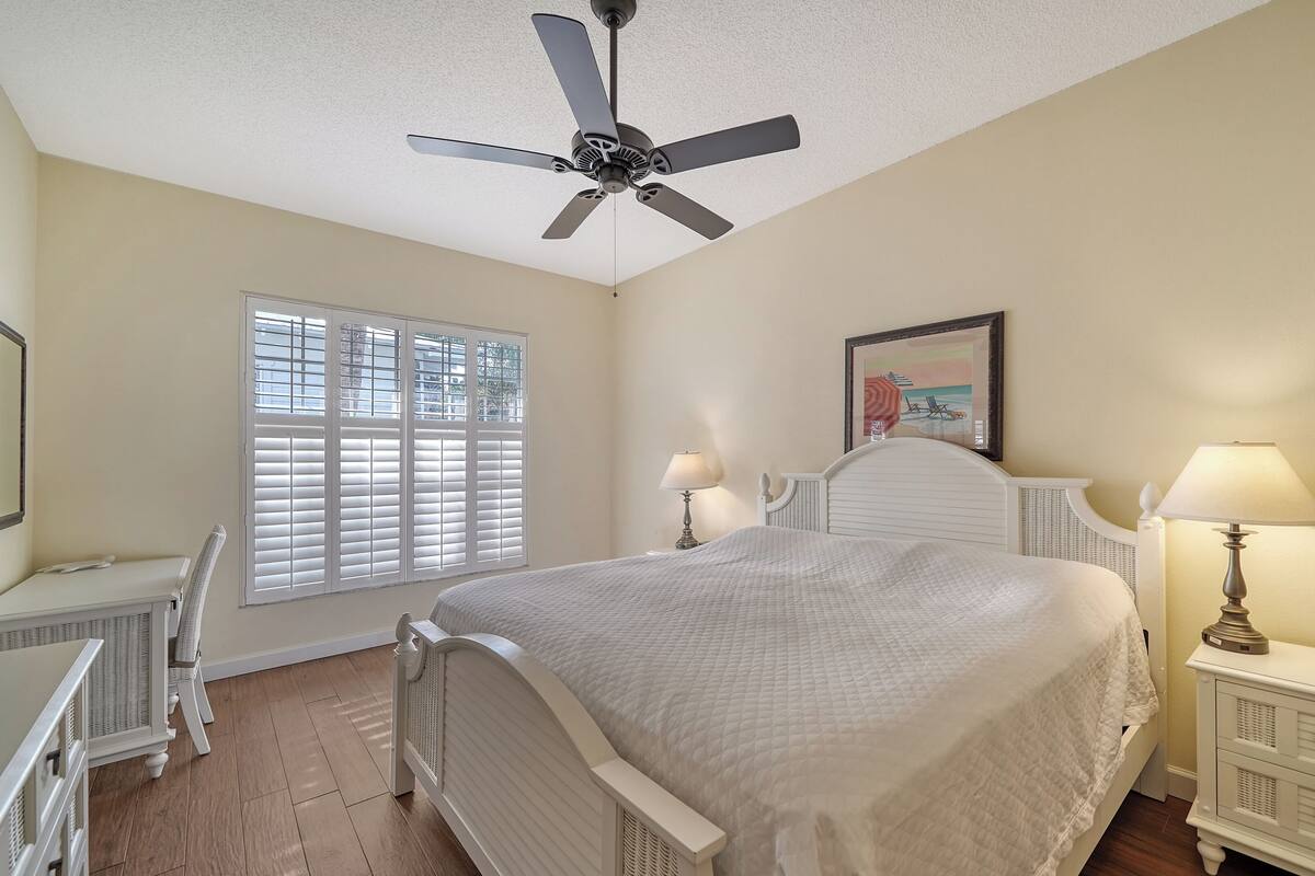 A bedroom with a bed, two bedside tables with lamps, a ceiling fan, a window with blinds, and a framed picture on the wall.