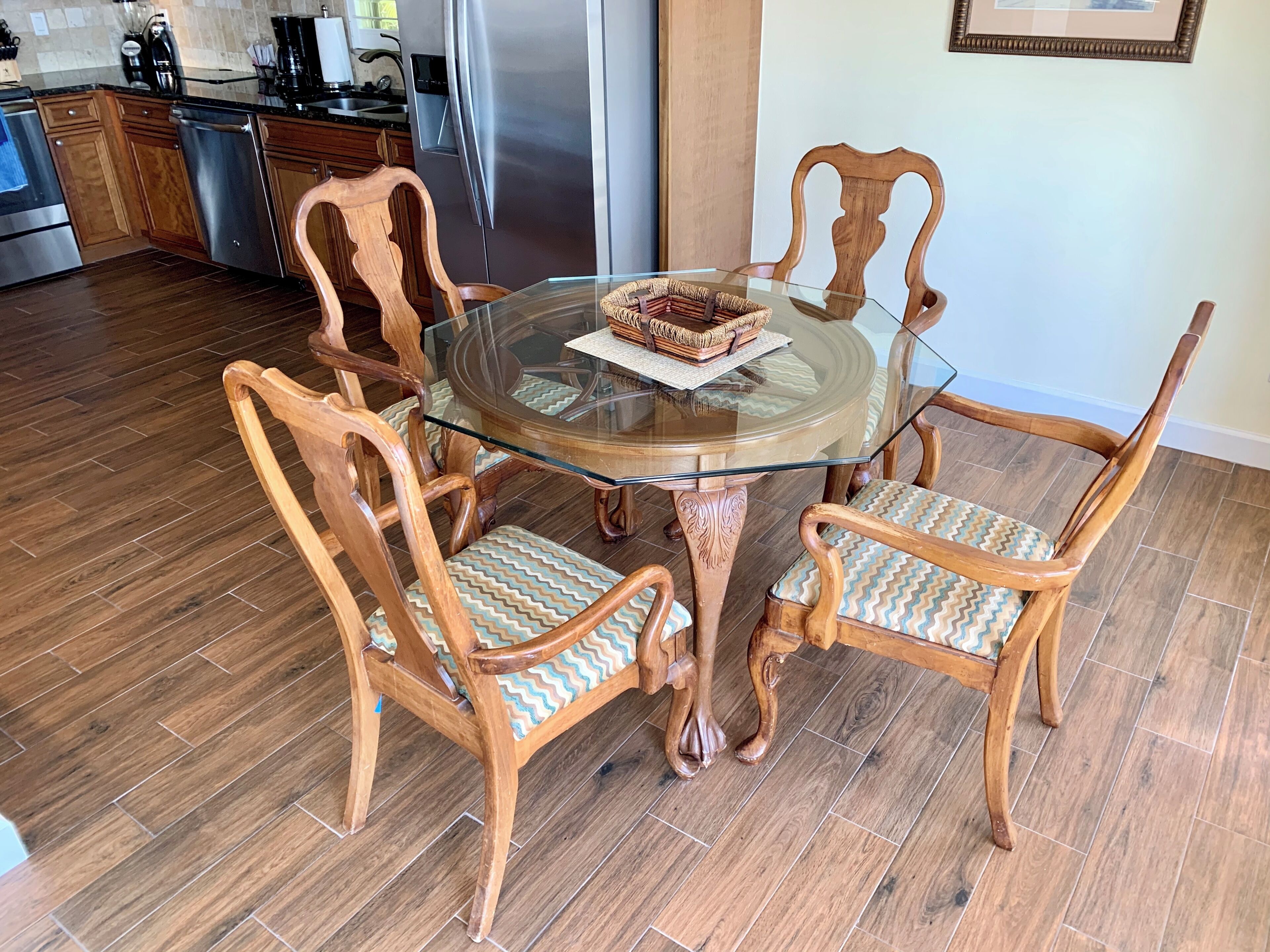 A round glass table with wooden chairs, a kitchen in the background, and a framed picture on the wall.