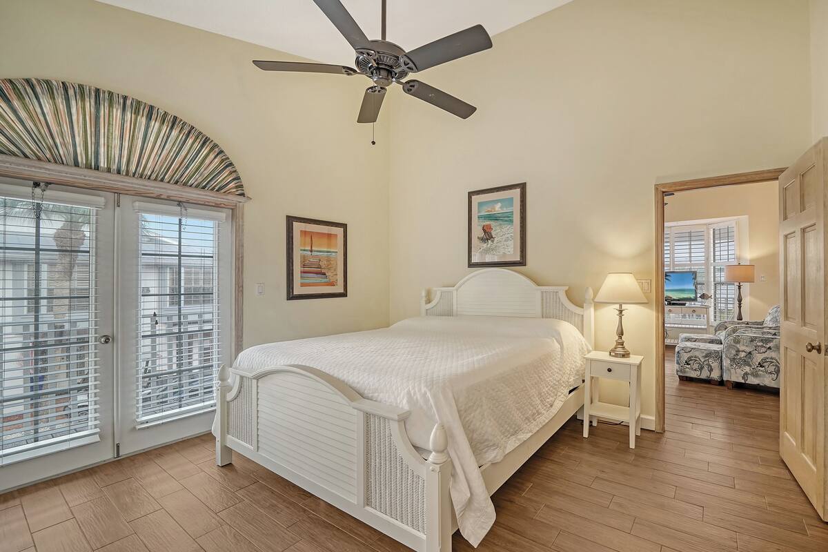 A bedroom with a bed, a ceiling fan, a nightstand, a lamp, a picture frame, and a view of another room through an open door.