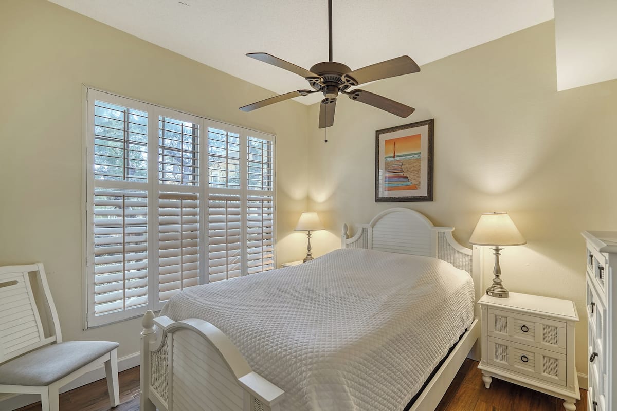 A bedroom with a bed, two bedside tables with lamps, a ceiling fan, a window with blinds, a chair, and a TV on a dresser.