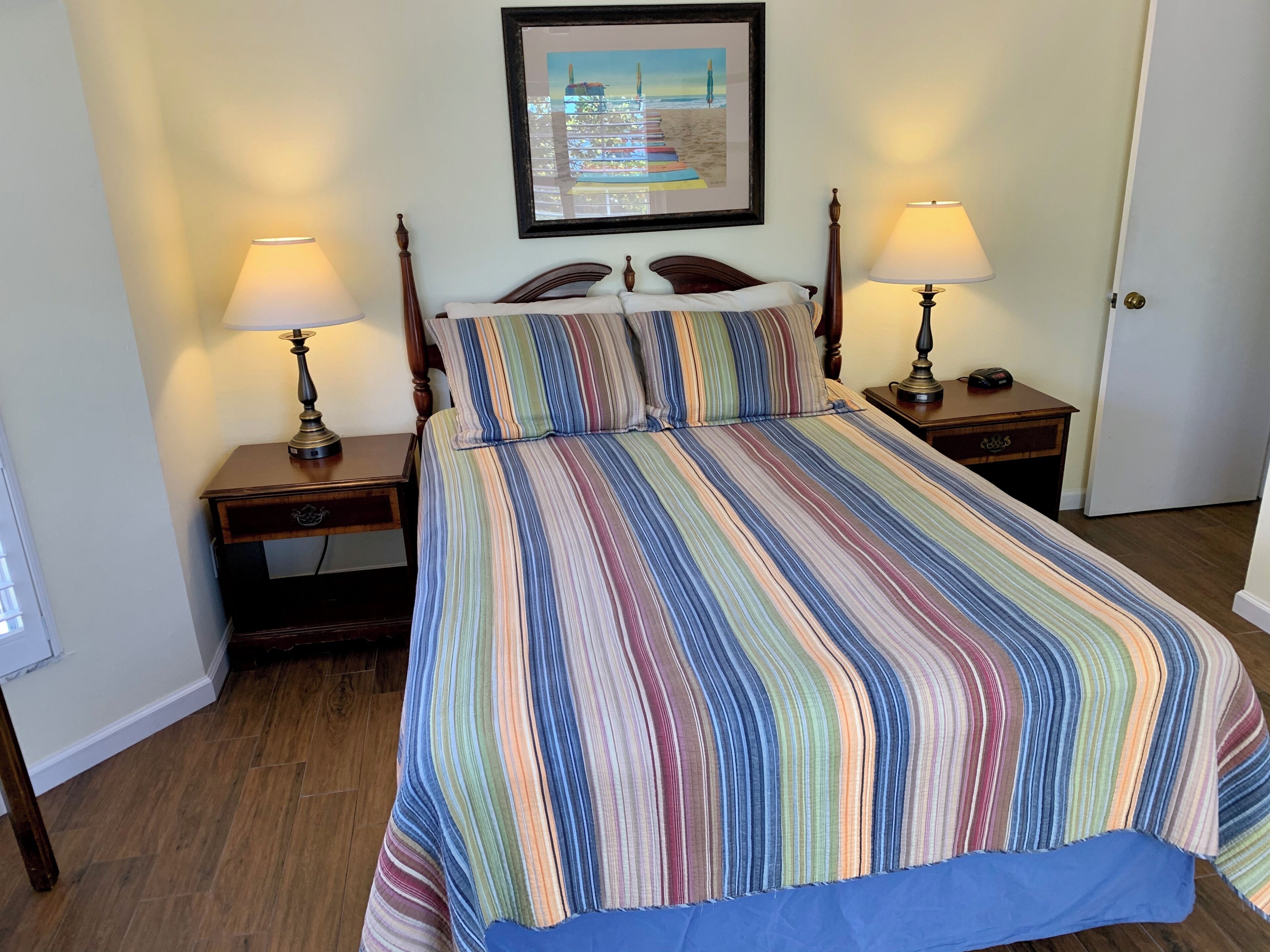 Una habitación de hotel con una cama, dos mesitas de noche, una lámpara de noche, un cuadro enmarcado en la pared y una puerta.