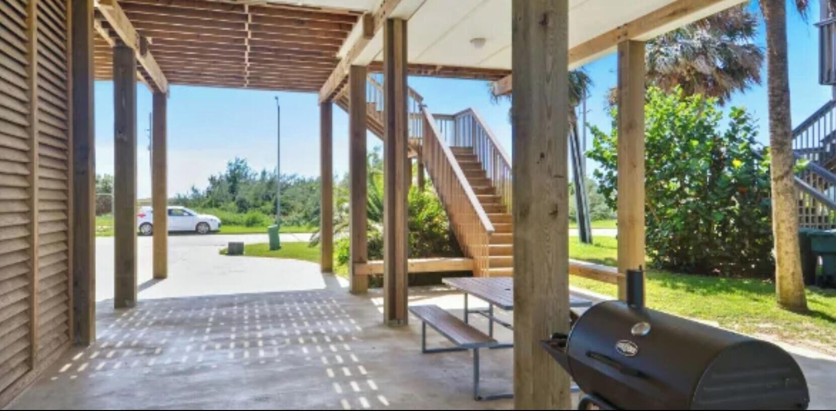 A covered outdoor area with a wooden staircase, picnic tables, and a barbecue grill.