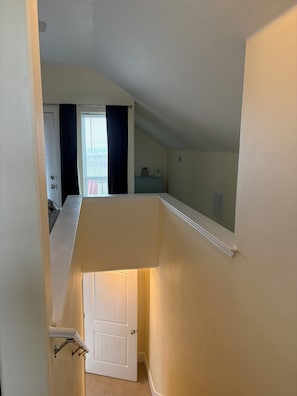 Eine enge Treppe mit einer Tür am unteren Ende, die zu einem Zimmer mit Fenster und Heizkörper führt.