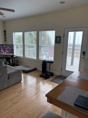 Ein Wohnzimmer mit grauem Sofa, einem Holztisch, einem Fernseher und einem großen Fenster mit Jalousien.