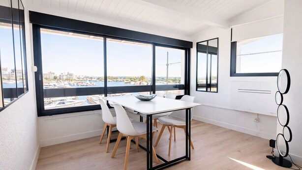 Una habitación moderna con una mesa de comedor, sillas, ventanas amplias y vistas a un puerto deportivo.