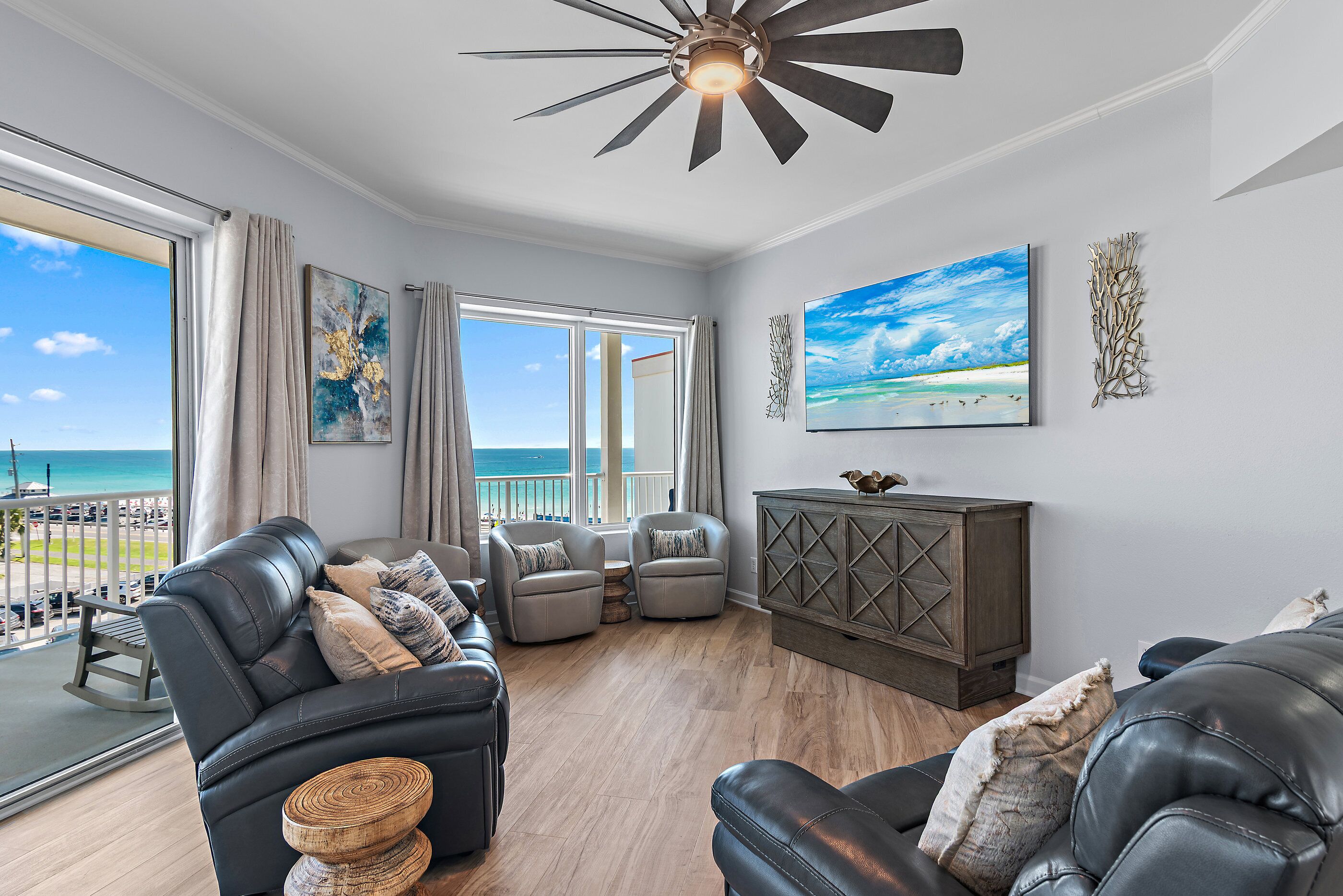 A spacious living room with a view of the beach, a ceiling fan, and a wooden cabinet.