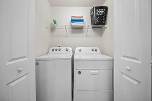 A laundry room with two white washing machines, a shelf with folded laundry, and a black laundry basket. - 5BR house sleeps 12 with game room and pool (Davenport)