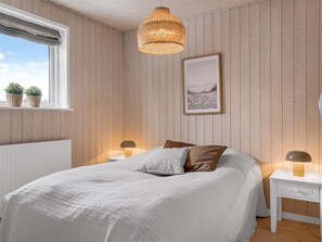 Une chambre à coucher avec un lit, des tables de chevet, un luminaire suspendu, une fenêtre avec des plantes et un tableau encadré au mur.