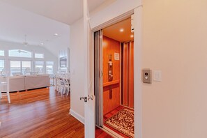 A spacious room with a large window, a dining area with chairs and a table, and a wooden door leading to another room. - JL #714 - Sunset Splendor -  Luxurious Soundfront Home - Hot tub - Elevator (Kill Devil Hills)