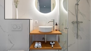 A modern bathroom with a round mirror, a white sink, a shower, and a wooden shelf with towels and a hairdryer.