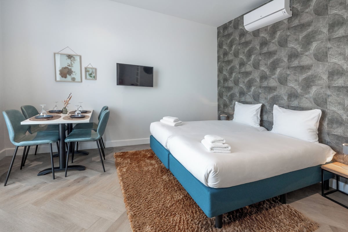 A modern hotel room with a bed, a dining table with chairs, a TV, and artwork on the wall.