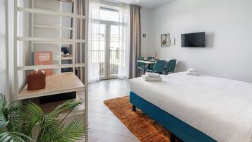 Habitación de hotel con cama, escritorio, silla, televisor y balcón con cortinas.