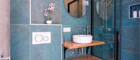 A bathroom with a glass shower enclosure, a white sink, and a wooden shelf.