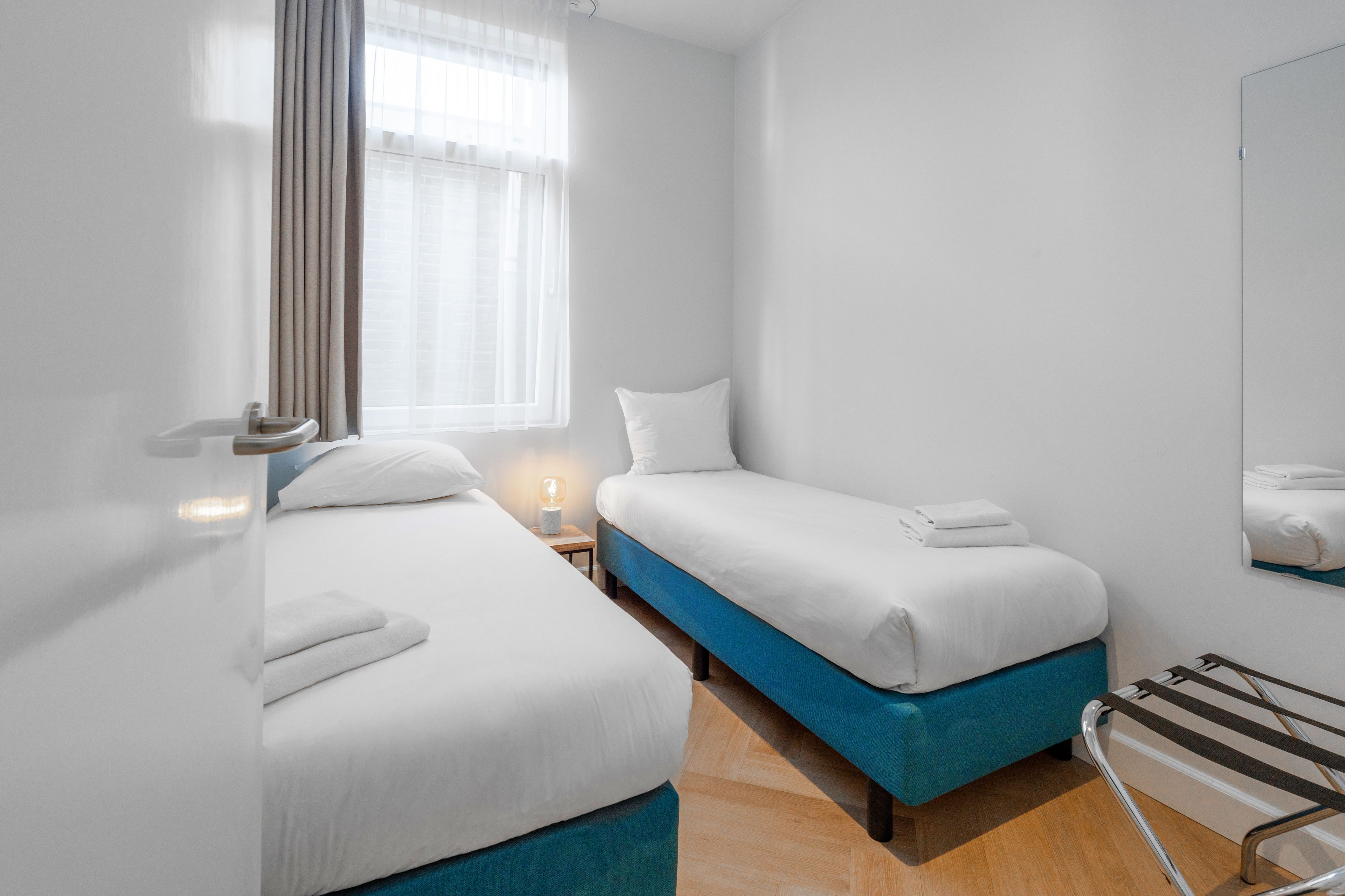 Une chambre d’hôtel avec deux lits, une fenêtre avec des rideaux, une table de chevet avec une lampe et un porte-serviettes.