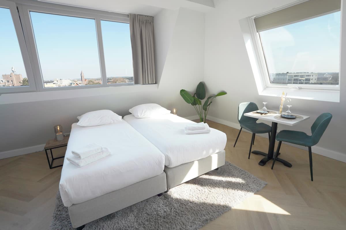 A bedroom with two beds, a desk, chairs, a plant, and a view of the city.