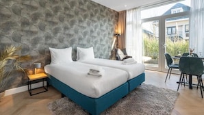 A modern bedroom with a large bed, a nightstand, a lamp, and a view of the outdoors.