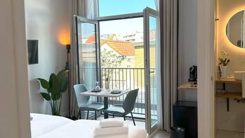 Une chambre d’hôtel comprenant un lit, une table à manger avec des chaises, un petit balcon, une salle de bain avec un miroir et une poubelle.