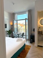 A hotel room with a bed, a dining table with chairs, a small balcony, a bathroom with a mirror, and a trash bin.