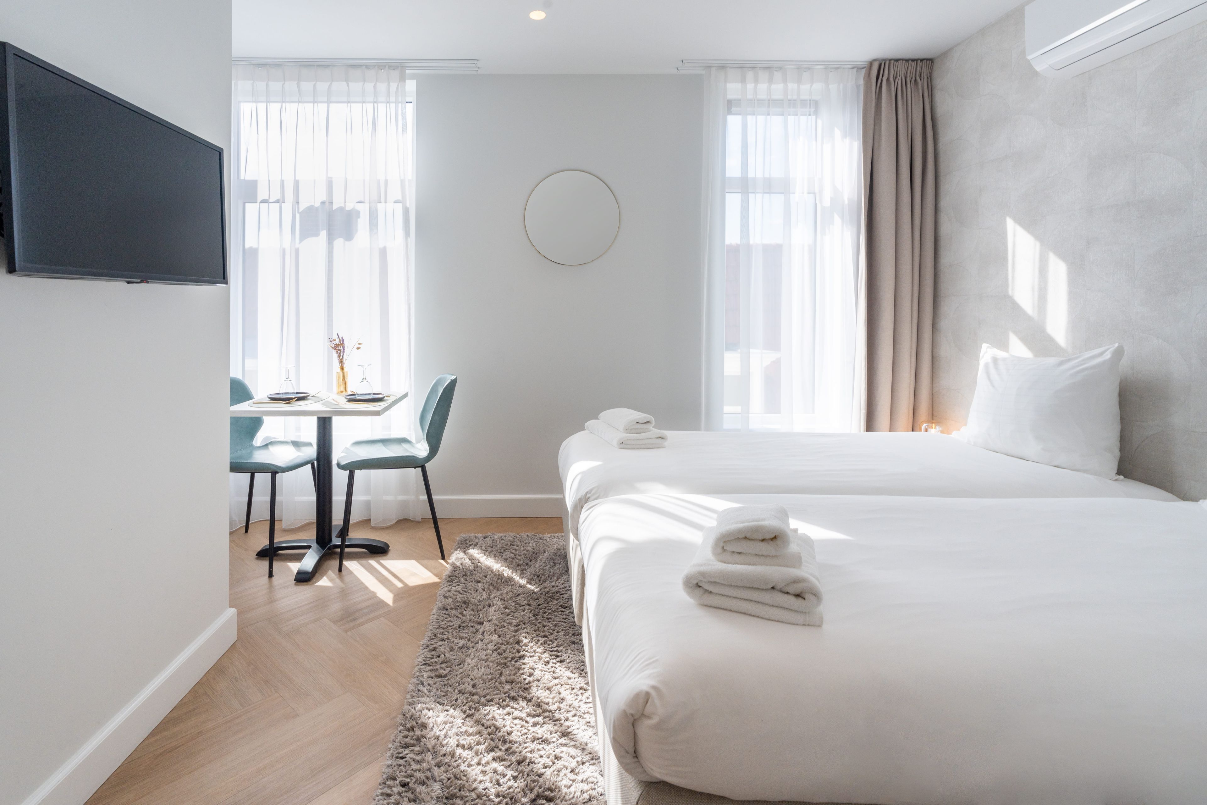 A hotel room with a bed, a desk with chairs, a TV mounted on the wall, and a window with curtains.