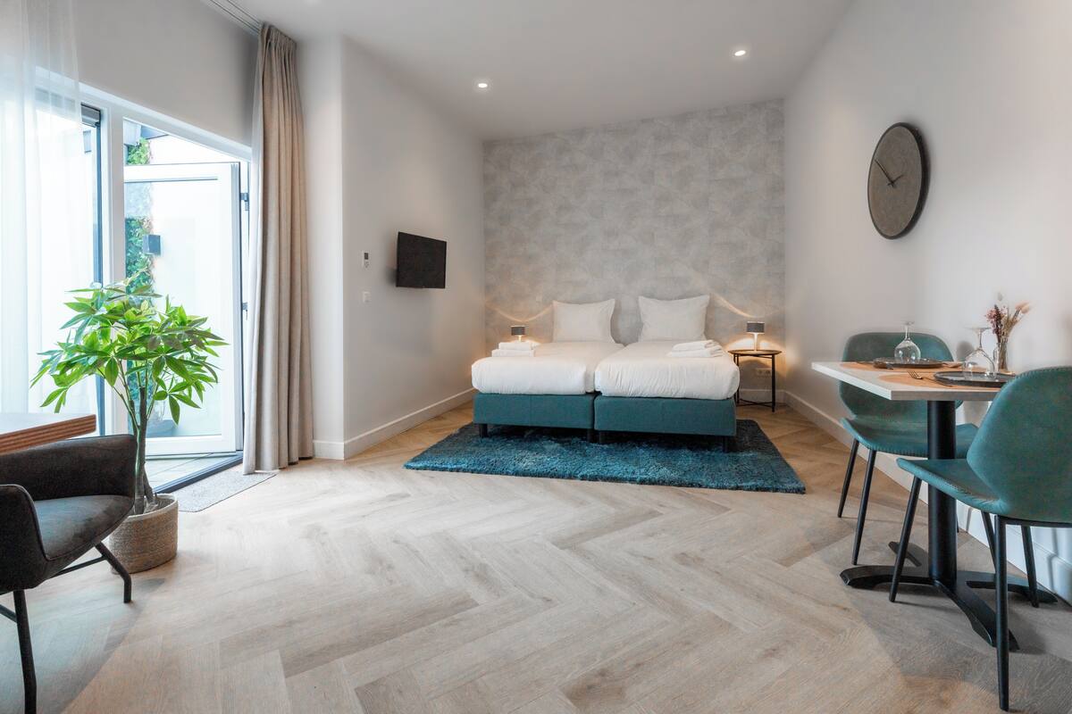 A modern hotel room with a bed, a dining table, a chair, a potted plant, and a view of the outdoors.