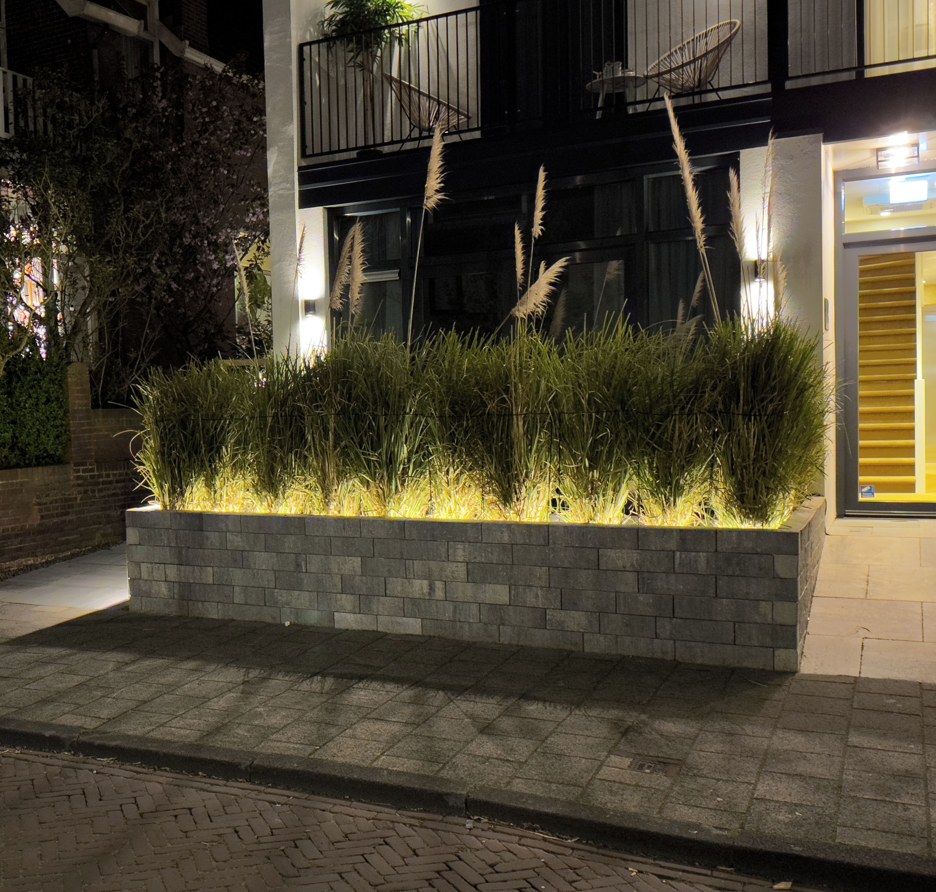 Vista nocturna de un edificio con jardín ajardinado, césped alto y maceta de ladrillo.