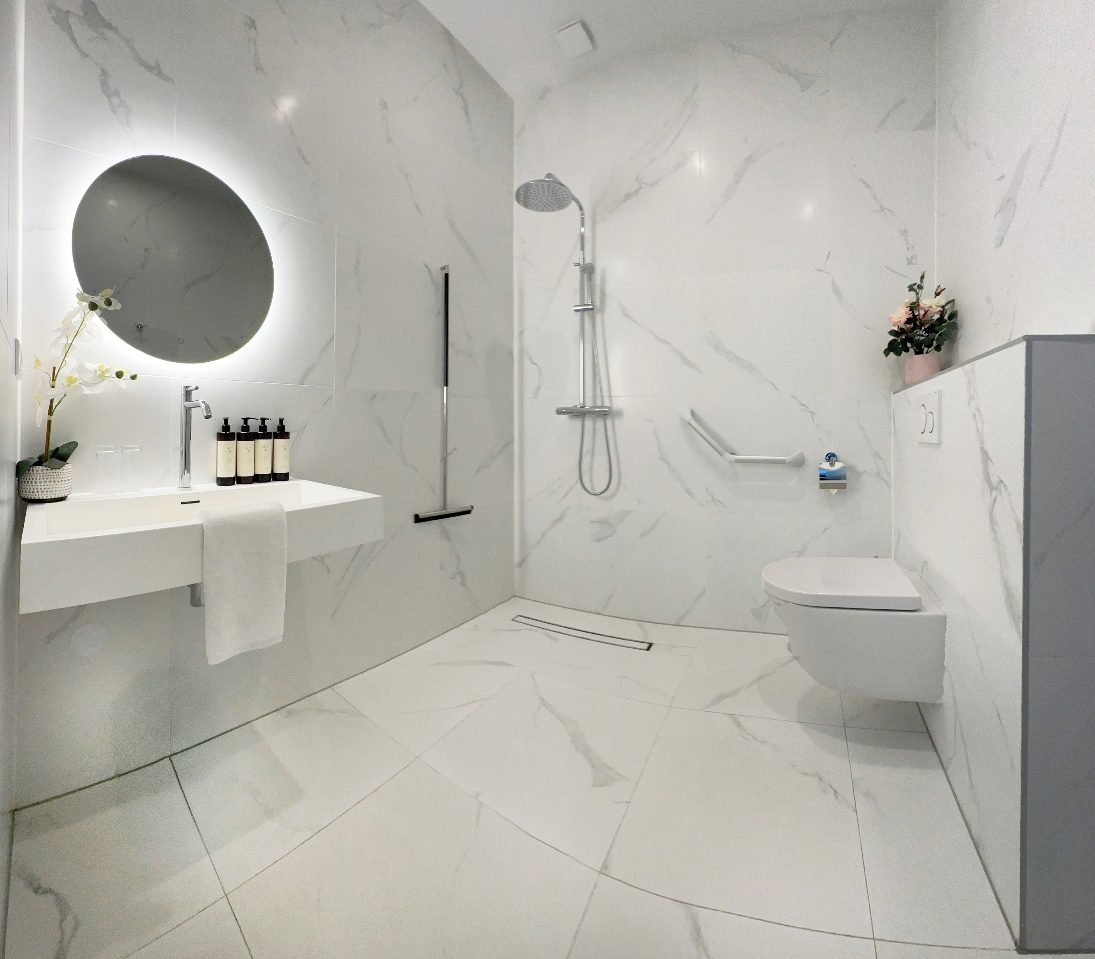 A modern bathroom with a white marble sink, a round mirror, a handheld showerhead, and a toilet.