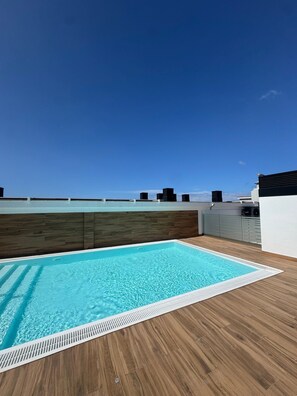 Una piscina en la azotea con cubierta de madera, cielo azul despejado y el horizonte de la ciudad al fondo.