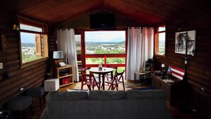 Ein gemütliches Blockhaus mit Holzvertäfelung, einem Essbereich mit roten Stühlen, einem an der Wand befestigten Fernseher und Blick auf eine grüne Landschaft durch große Fenster.