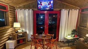 Ein gemütliches Blockhäuschen-Interieur mit einem Holzespeisetisch, Stühlen, einem an der Decke befestigten Fernseher und Blick auf die beleuchtete Stadt durch die Glastür.