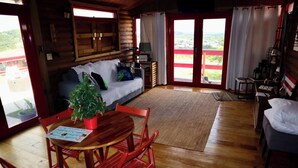 Ein Holzblockhaus-Interieur mit einem Holzesstisch, roten Stühlen, einer Couch und einem Weihnachtsbaum, sowie Blick durch Glastüren nach draußen.