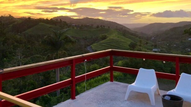 Ein Balkon mit zwei weißen Stühlen und einem roten Geländer mit Blick auf eine hügelige Landschaft bei Sonnenuntergang.
