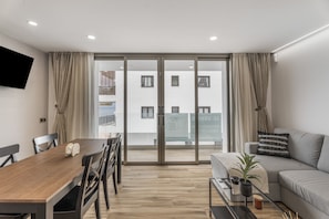 Un salon moderne comprenant un coin repas, un canapé, un téléviseur fixé au mur et un balcon offrant une vue sur les immebles.