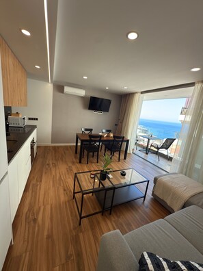 Ein modernes Wohnzimmer mit Holzboden, einem Glastisch, einer Couch, einem Essbereich mit Stühlen, einem an der Wand montierten Fernseher und Meerblick.