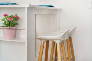 A white bar stool with wooden legs and a white seat, positioned next to a white shelf with a pink pot of flowers.