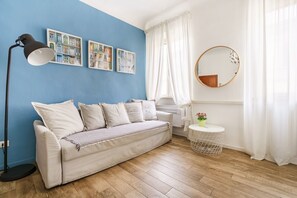 A room with a blue wall, a white sofa, a floor lamp, a round mirror, and a small round table with a plant.