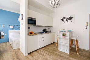 A modern kitchen with white cabinets, a microwave, and a sink. There's a dining area with a white table and a chair, and a bed with white bedding in the adjacent room.