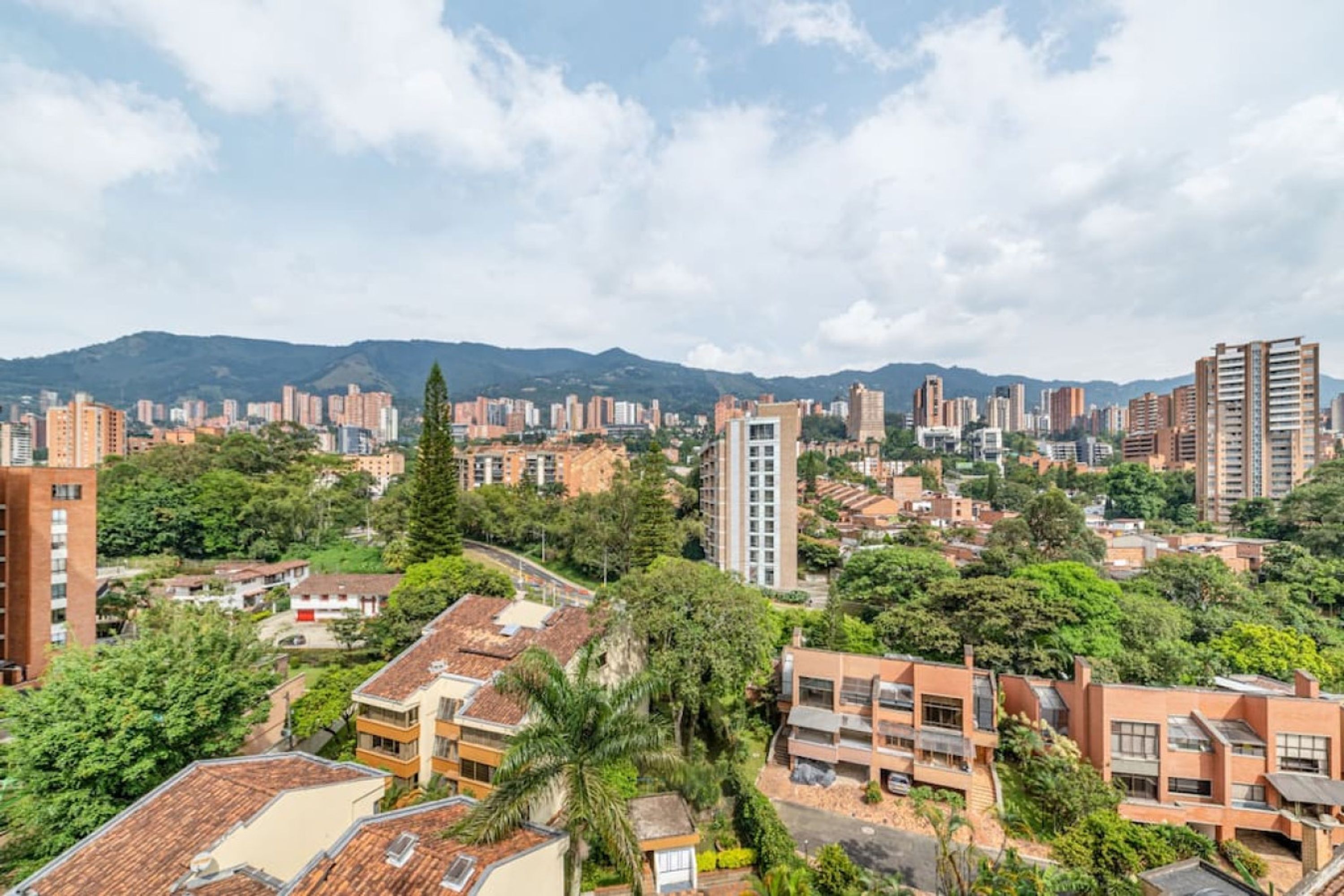 A cityscape with high-rise buildings, dense greenery, and a mountain range in the background.