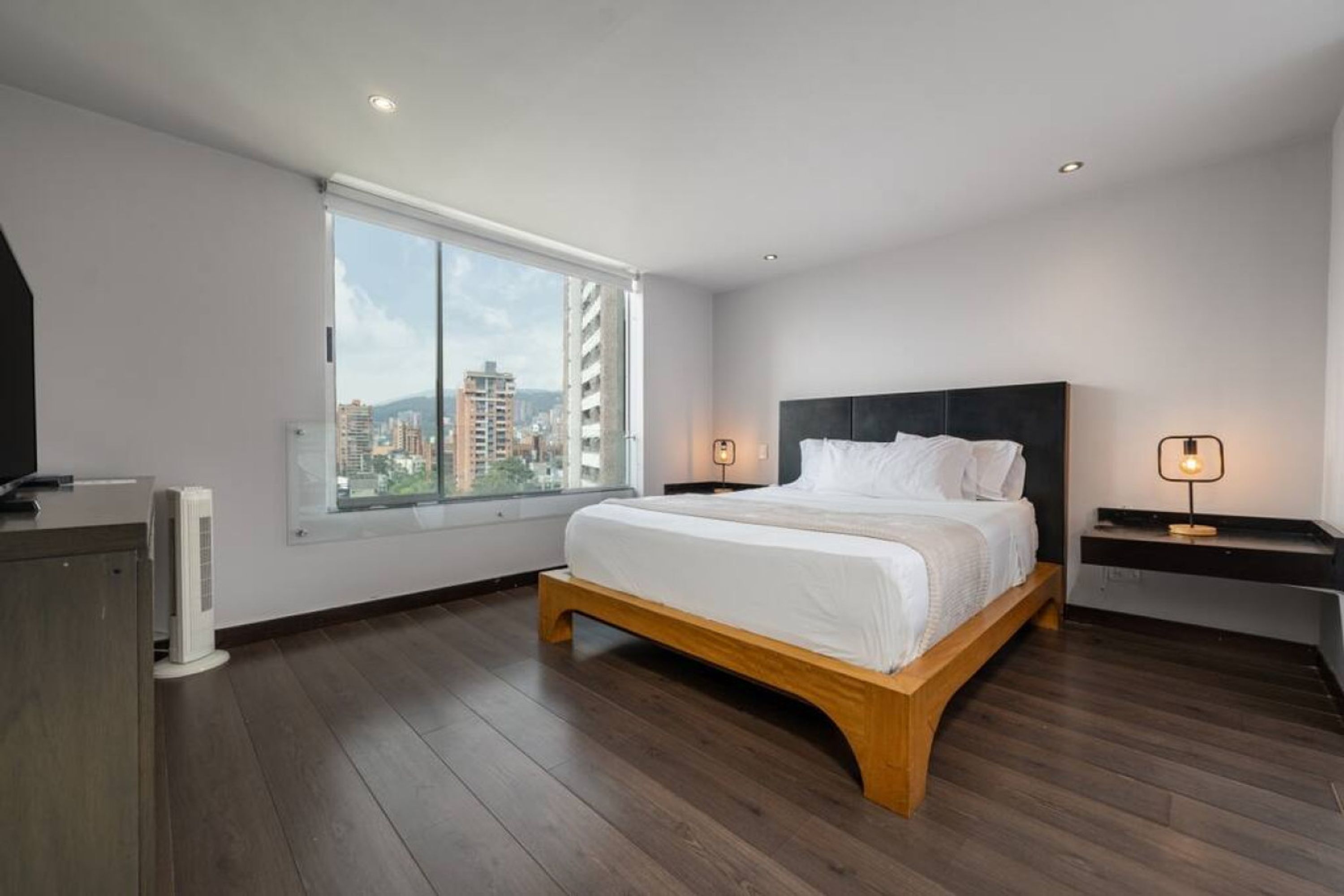 A modern bedroom with a large window, a wooden bed frame, and a nightstand with a lamp.