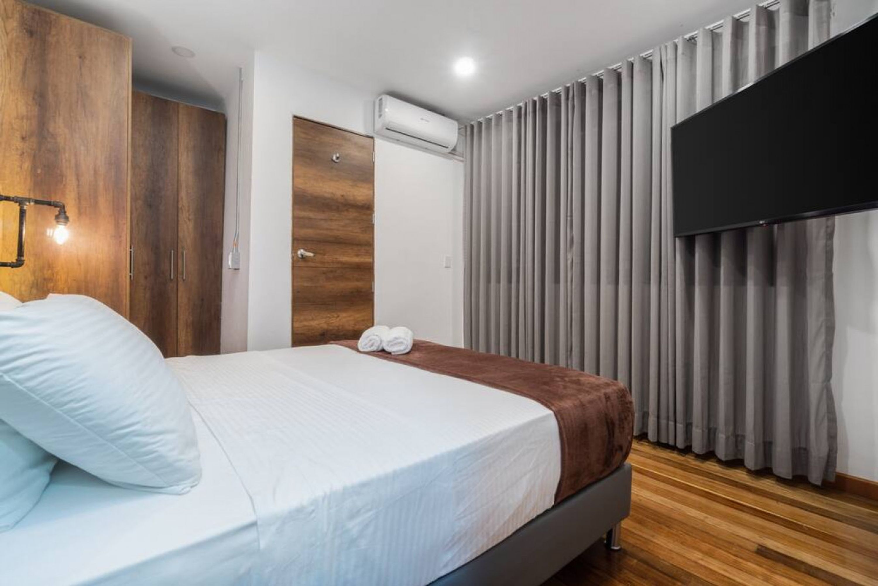 Quarto de hotel moderno com cama, guarda-roupa de madeira, ar condicionado e televisão montada na parede.