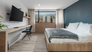 A modern hotel room with a large bed, a desk with a chair, a TV, and a city view. - Go Living Suites - 1305 (Medellín)