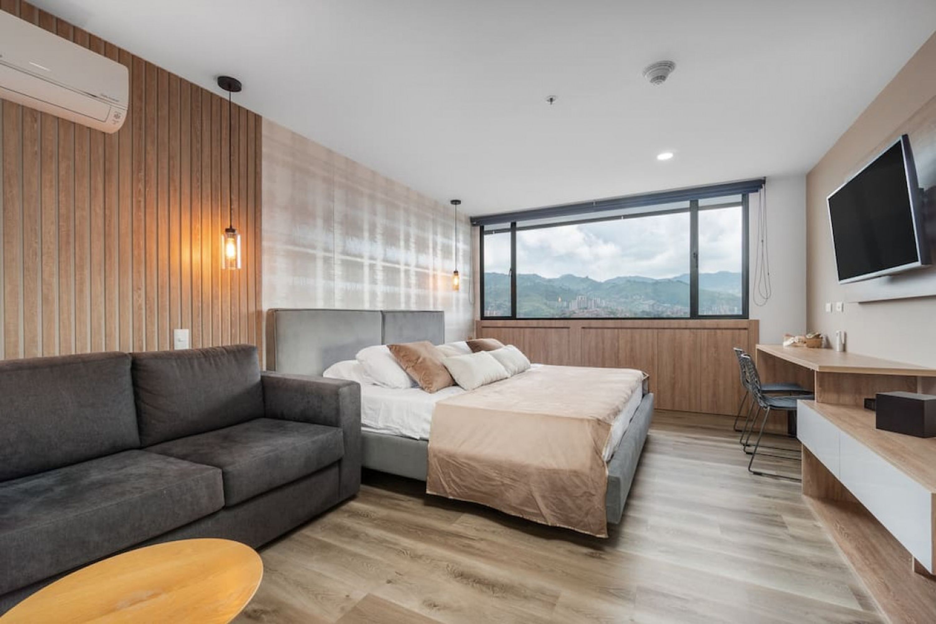 A modern hotel room with a bed, a sofa, a TV, and a large window with a mountain view.